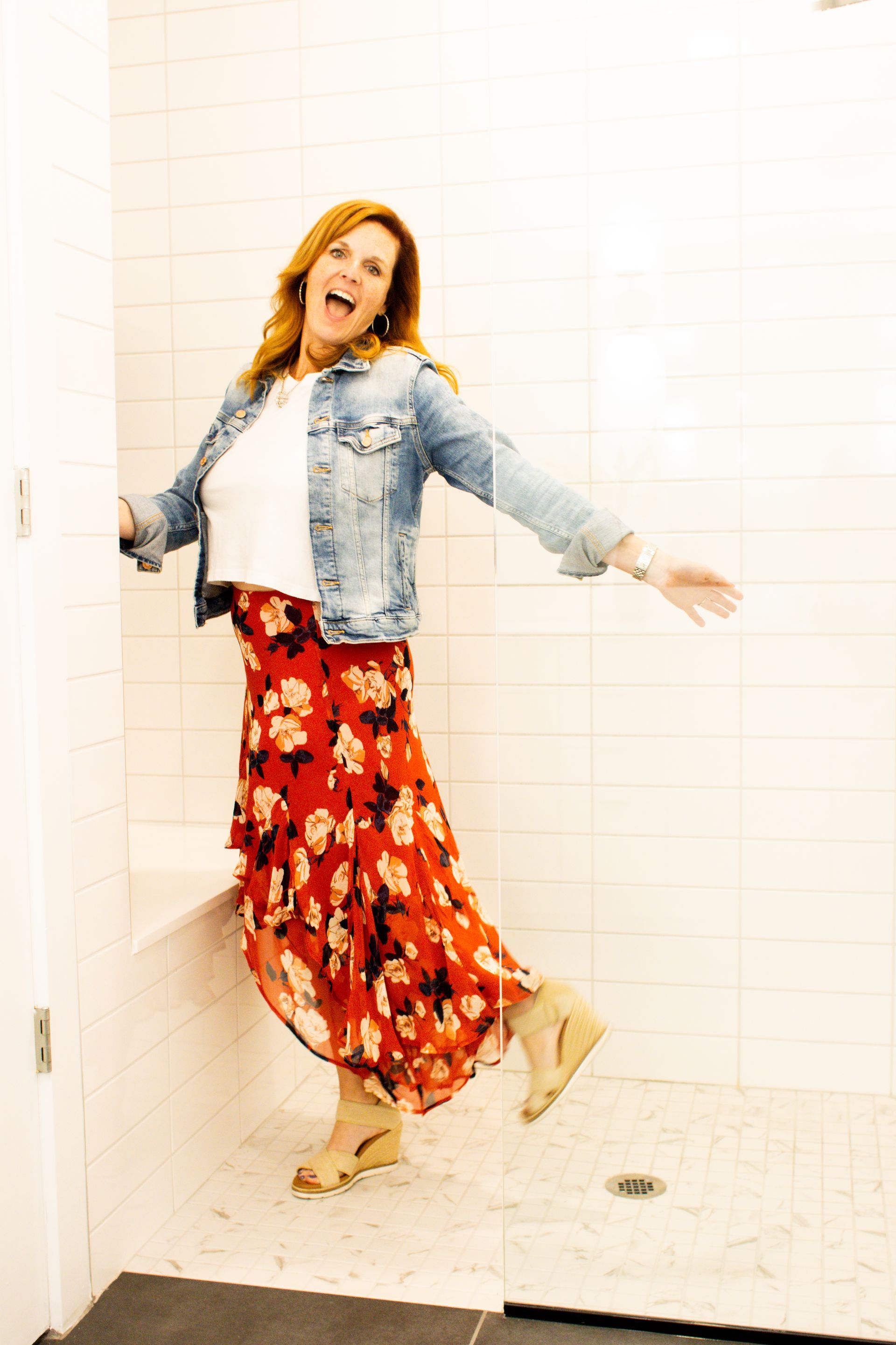 Woman in a denim jacket and floral skirt stands in a white-tiled shower, smiling with arms out.