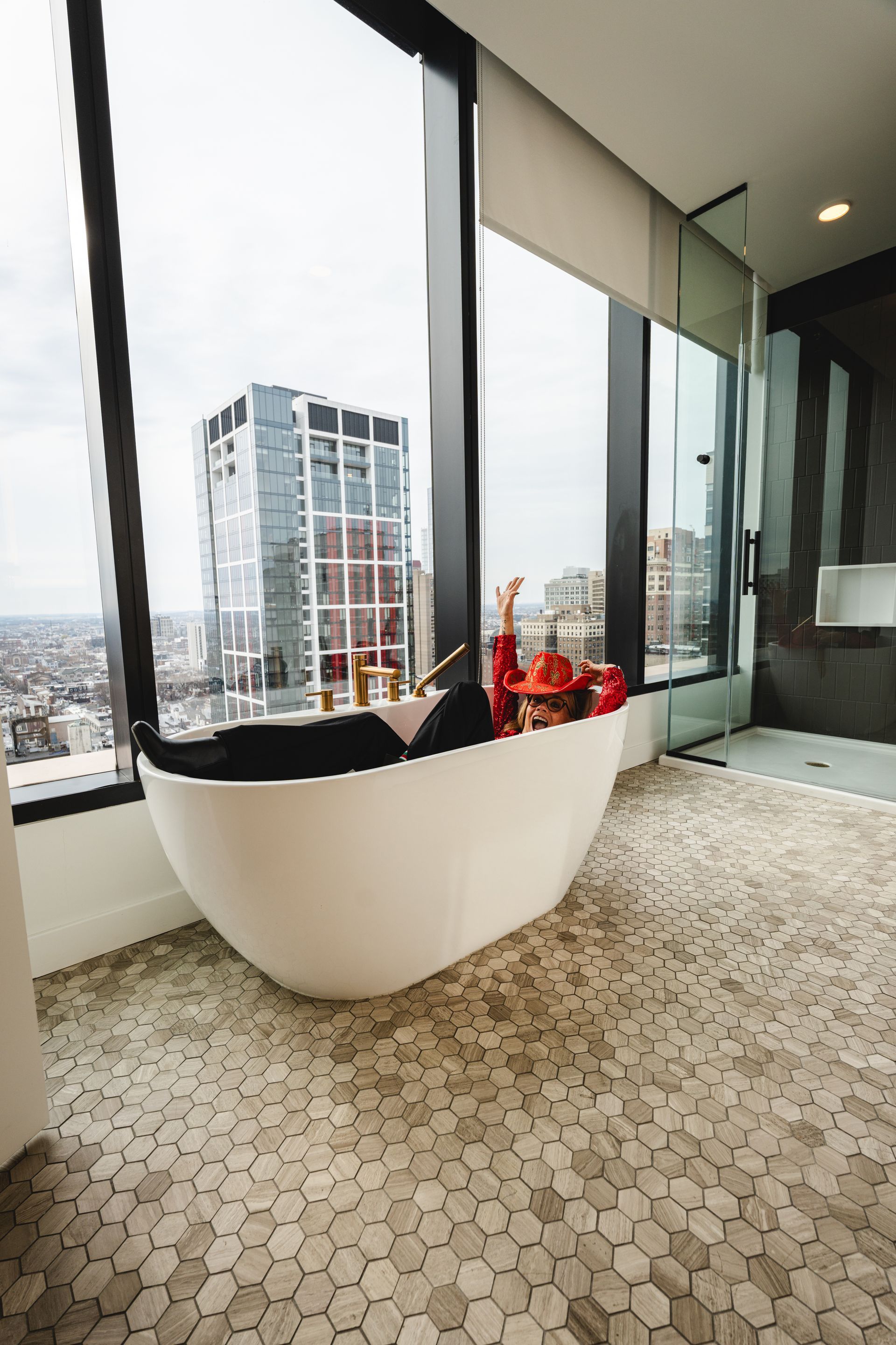 White oval bathtub near a tall window with city view, honeycomb tile floor.