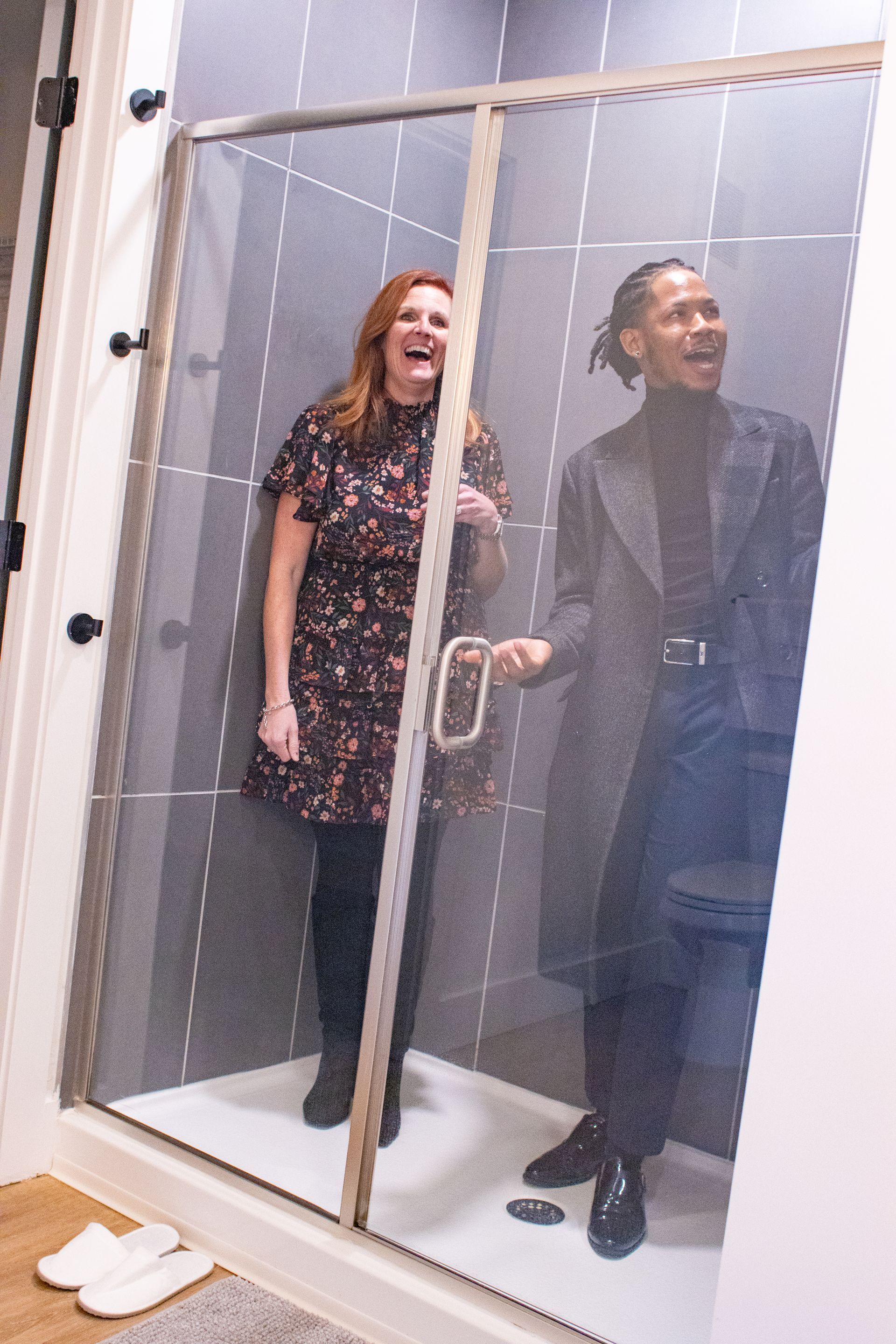Woman and man inside a glass shower stall, smiling. Woman wearing floral dress. Man wearing coat.