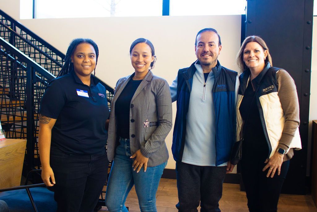 Four people smiling, posing together indoors near a staircase; neutral expressions; dressed casually.