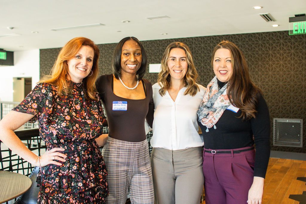 Four women smiling, posing indoors. One wears a floral dress, others wear business casual attire.
