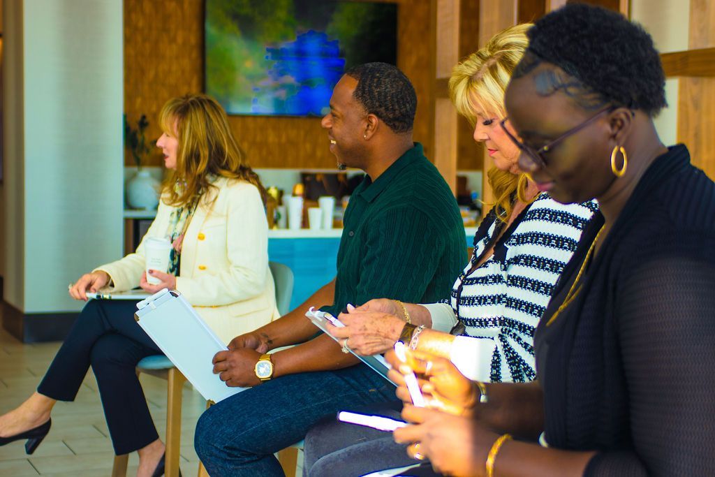 Four people seated, holding papers and pens, looking to the side, in a well-lit interior space.