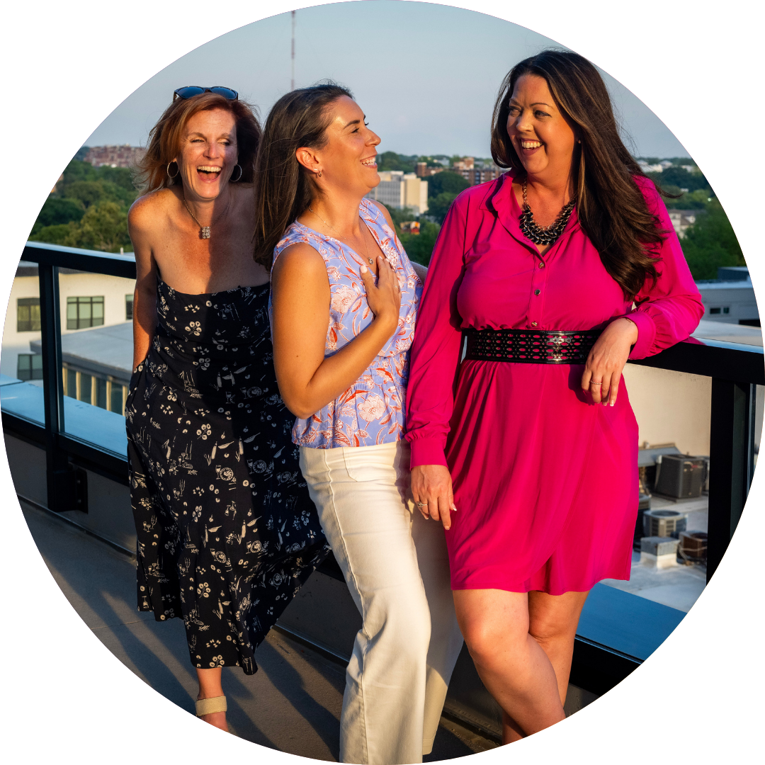 Three women laughing on a rooftop, leaning on a railing. Bright sun, city backdrop.
