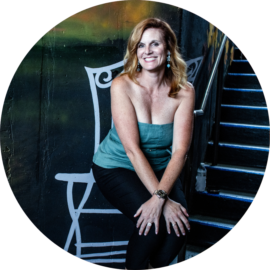 Woman in a green strapless top and black pants smiling, sitting by a white painted chair and stairs.