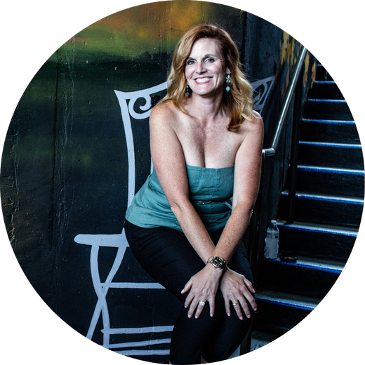 Woman in a green strapless top and black pants smiling, sitting by a white painted chair and stairs.