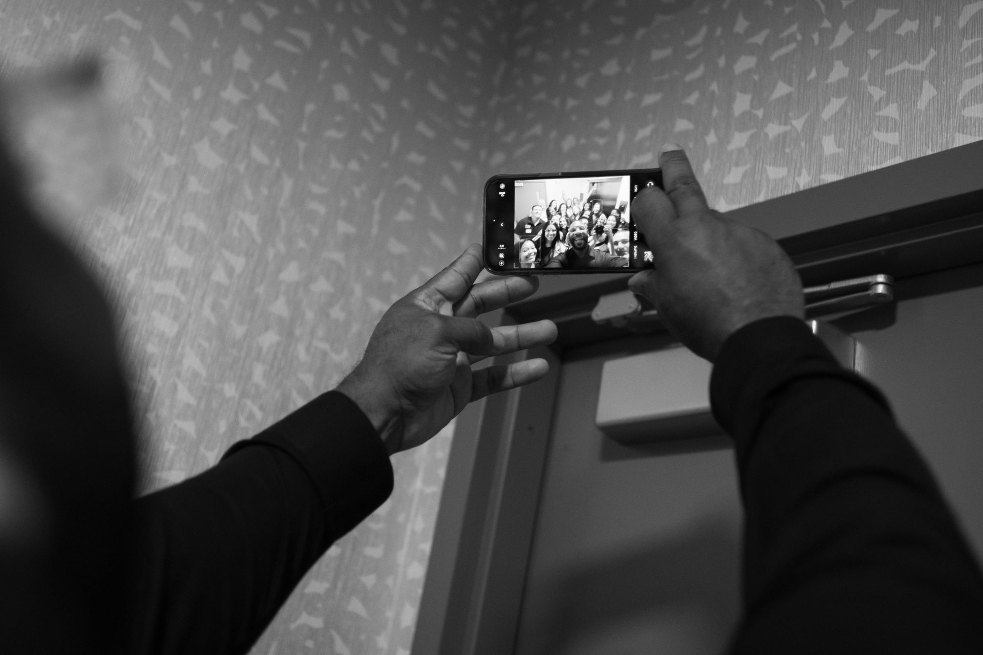 Hands holding a phone, filming a group through a doorway. Interior shot. Black and white.
