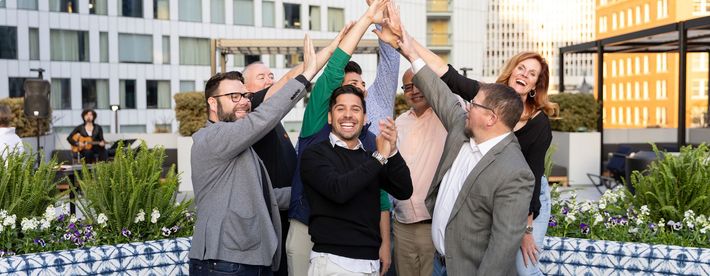 Group of people celebrating outdoors with hands raised. Buildings in the background.