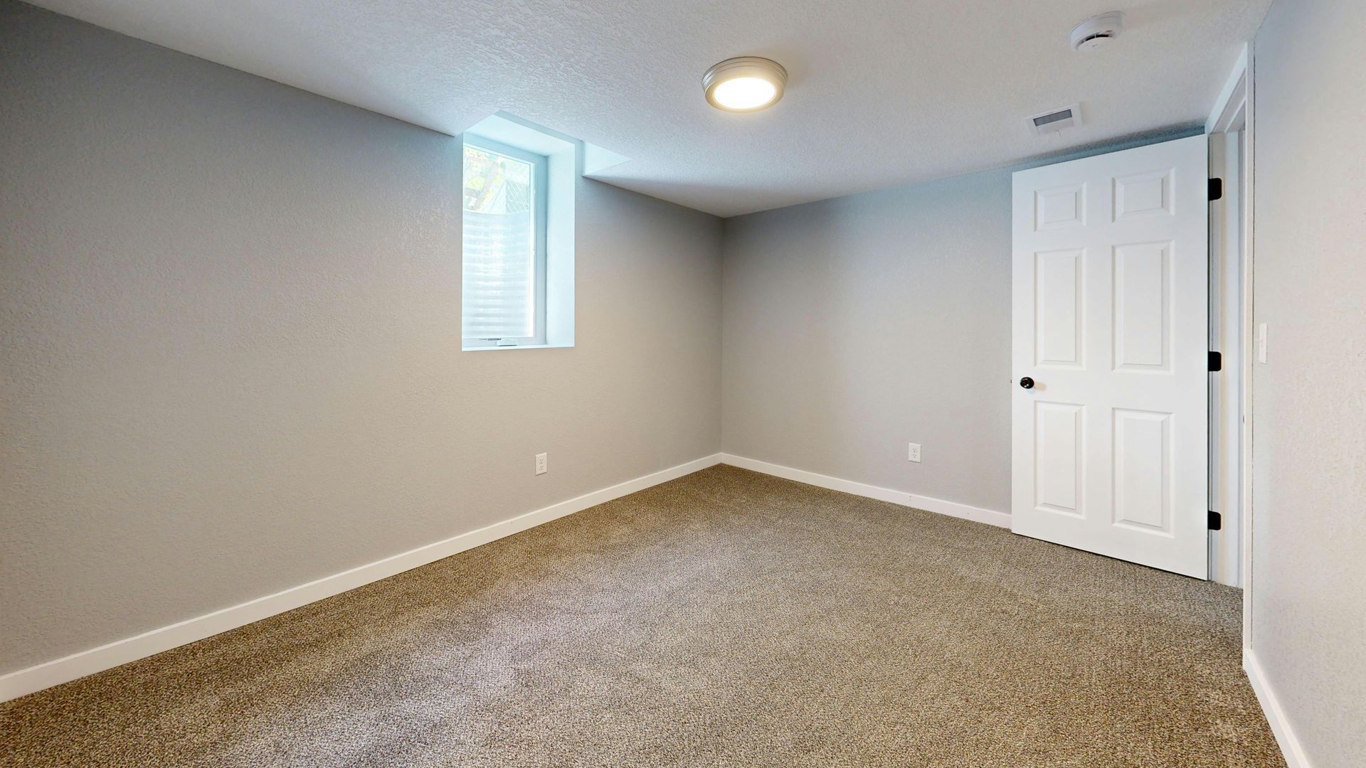 Empty beige bedroom with carpet, gray walls, small window, ceiling light, and white closet door