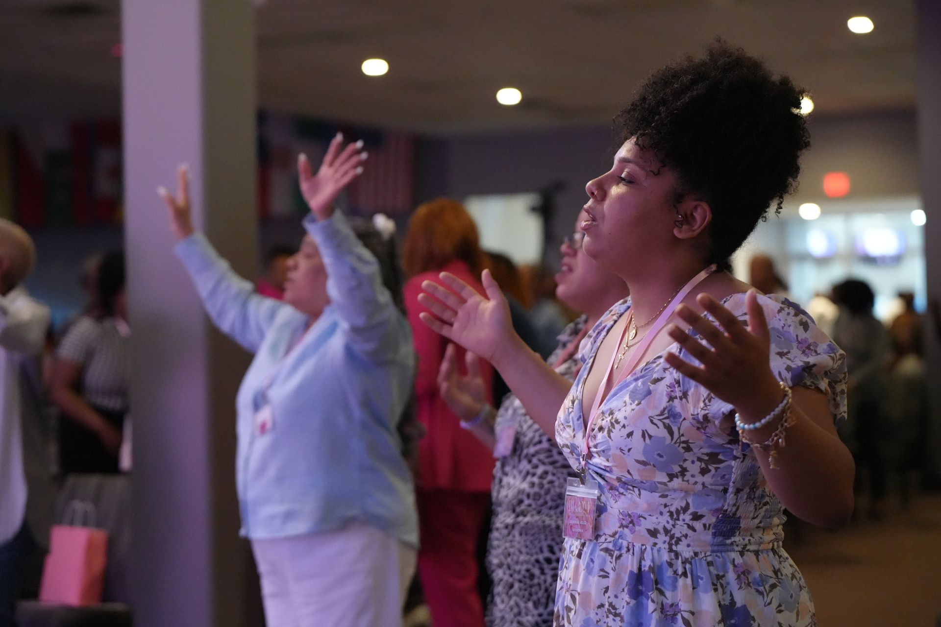 People in a light-filled space with arms raised. One woman sings with a floral dress and another in a light blue shirt.