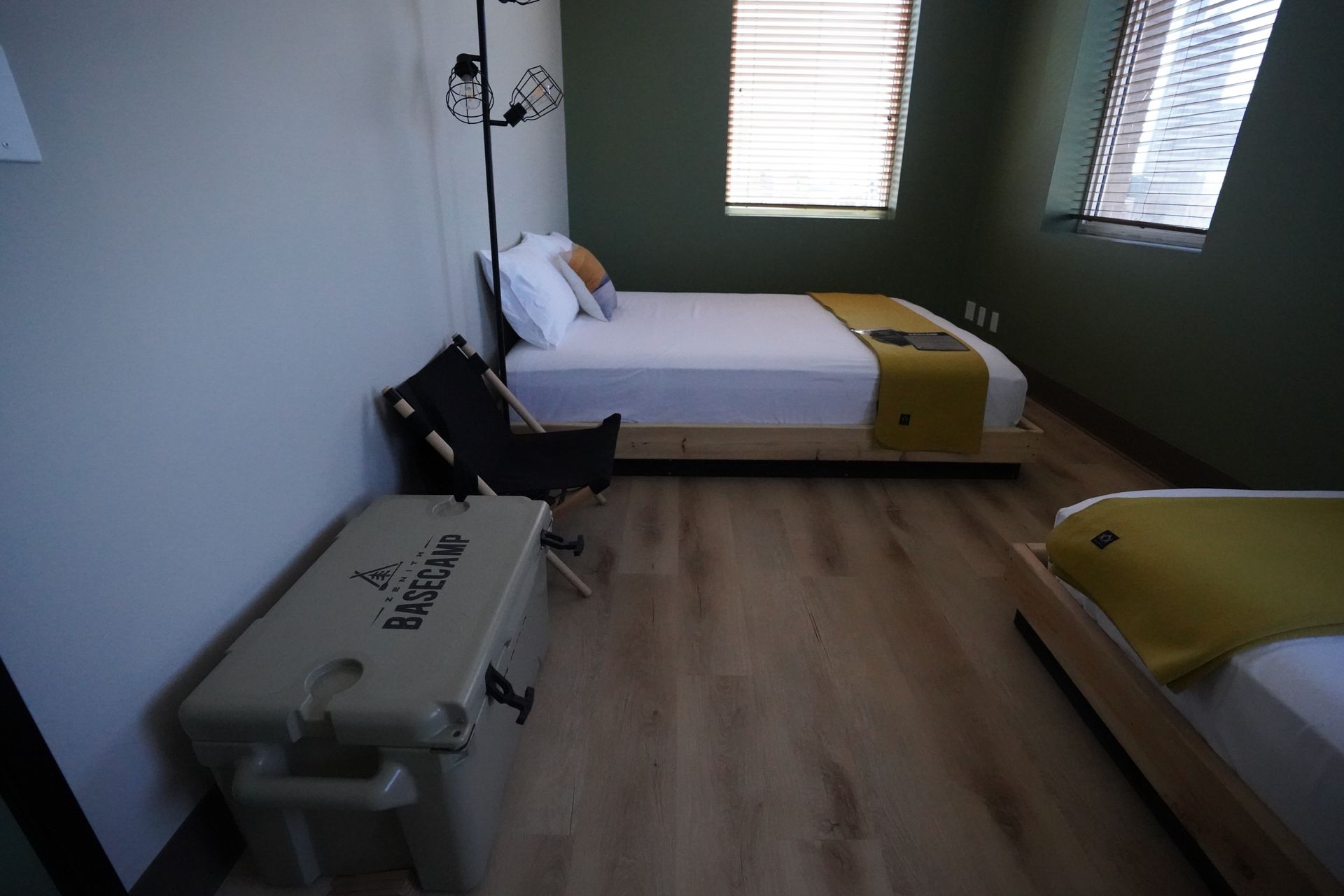 Bedroom with two beds, one chair, and a cooler. Light-colored wooden floors, dark green walls, and white bedding.