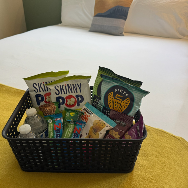 Basket of snacks, water bottles, on a yellow blanket, in front of a white bed with pillows.