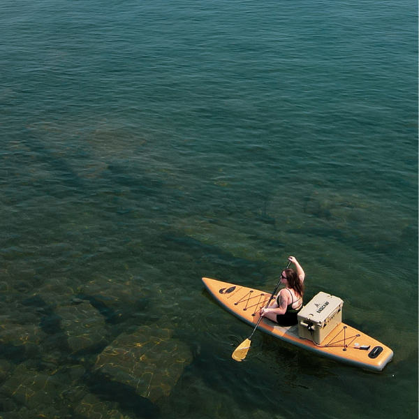 Woman on yellow paddleboard in clear water, arm raised; cooler present.