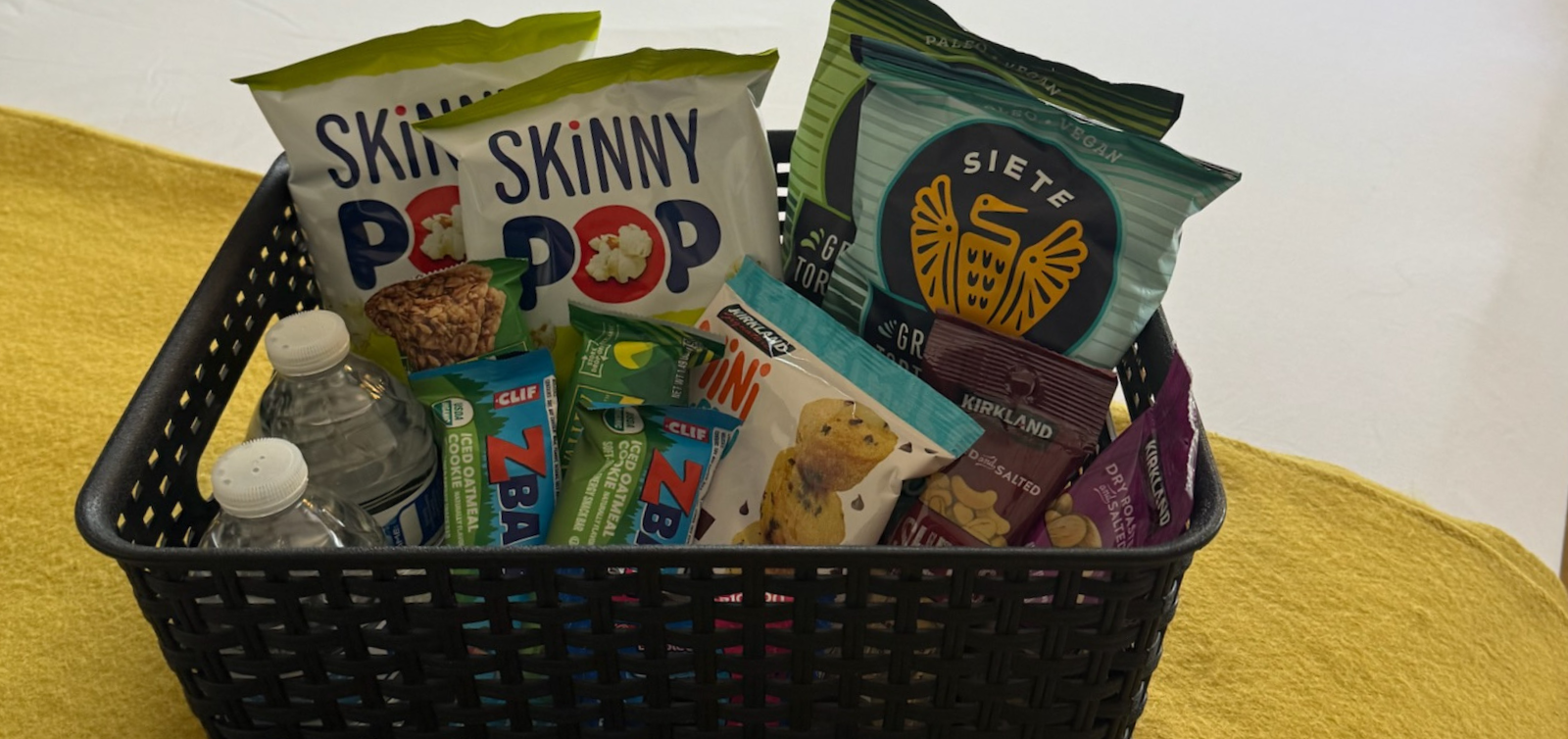 A black basket filled with snacks and water bottles on a yellow surface.
