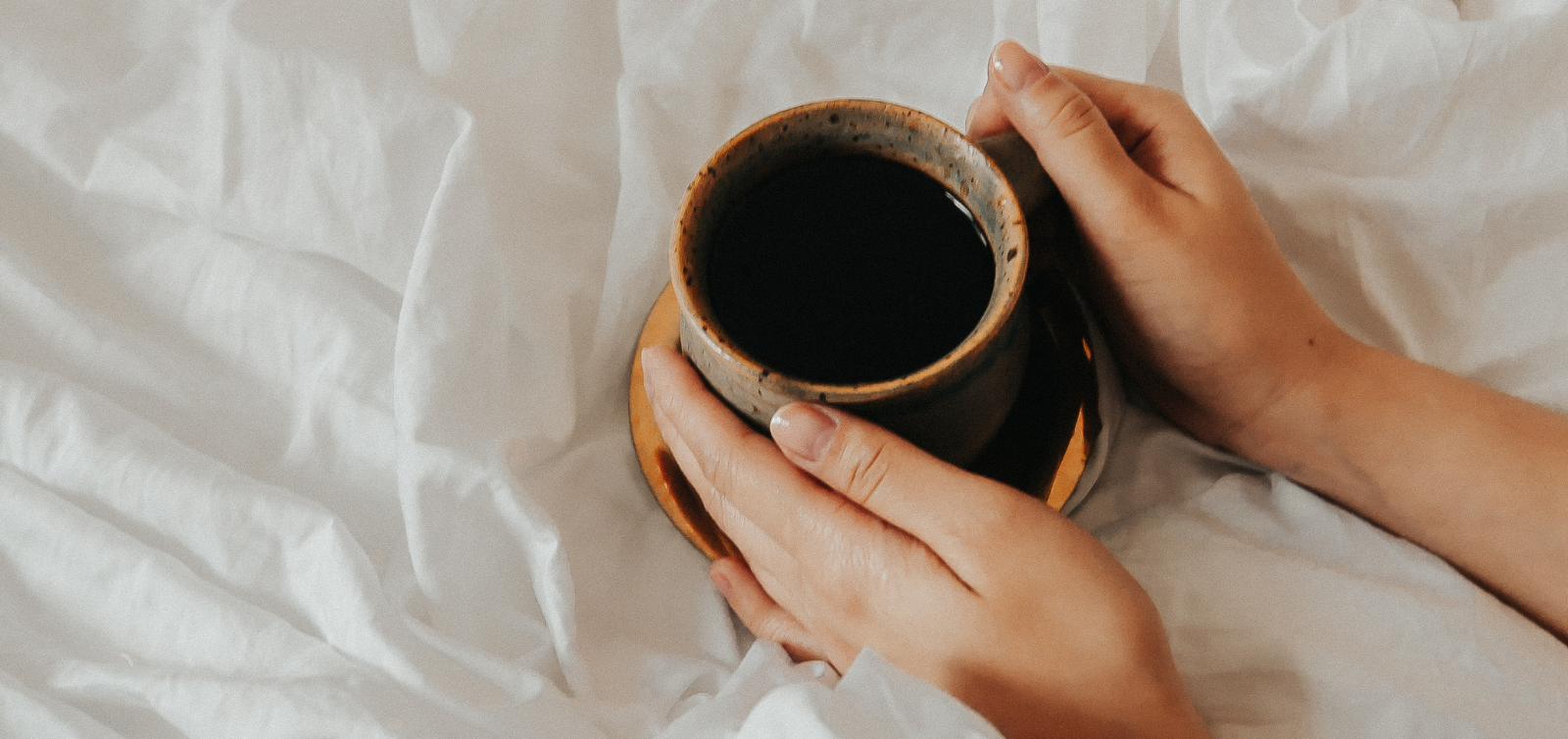 Person holding a mug of dark coffee on a white bed.