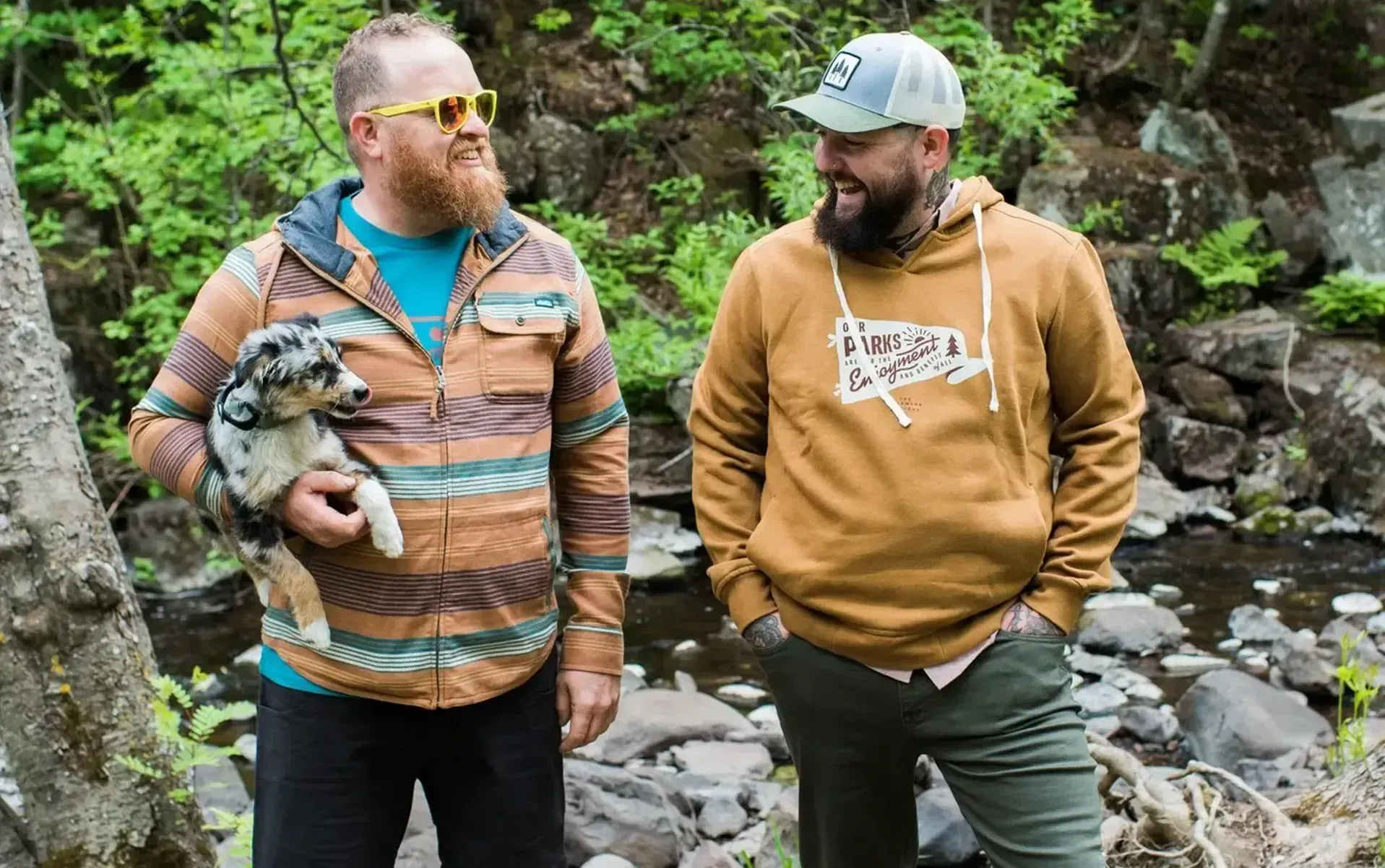 Two men looking and laughing at each other standing outside. One is holding a small dog.