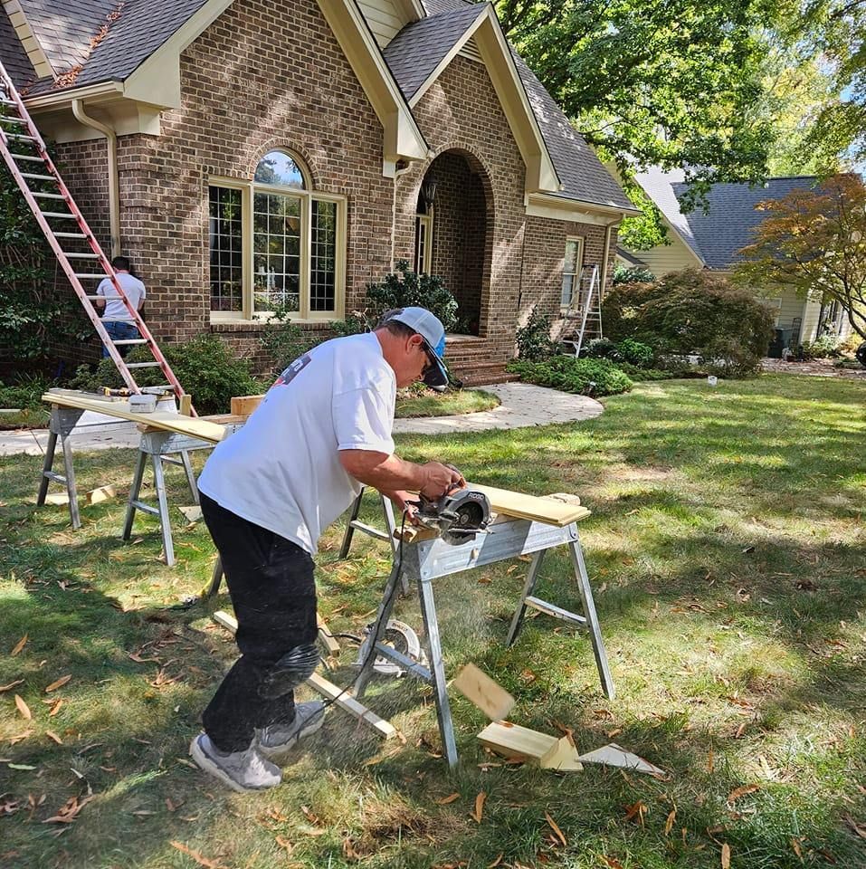Man using a circular saw on wood, outside a brick house.  Another person on a ladder. Green grass.