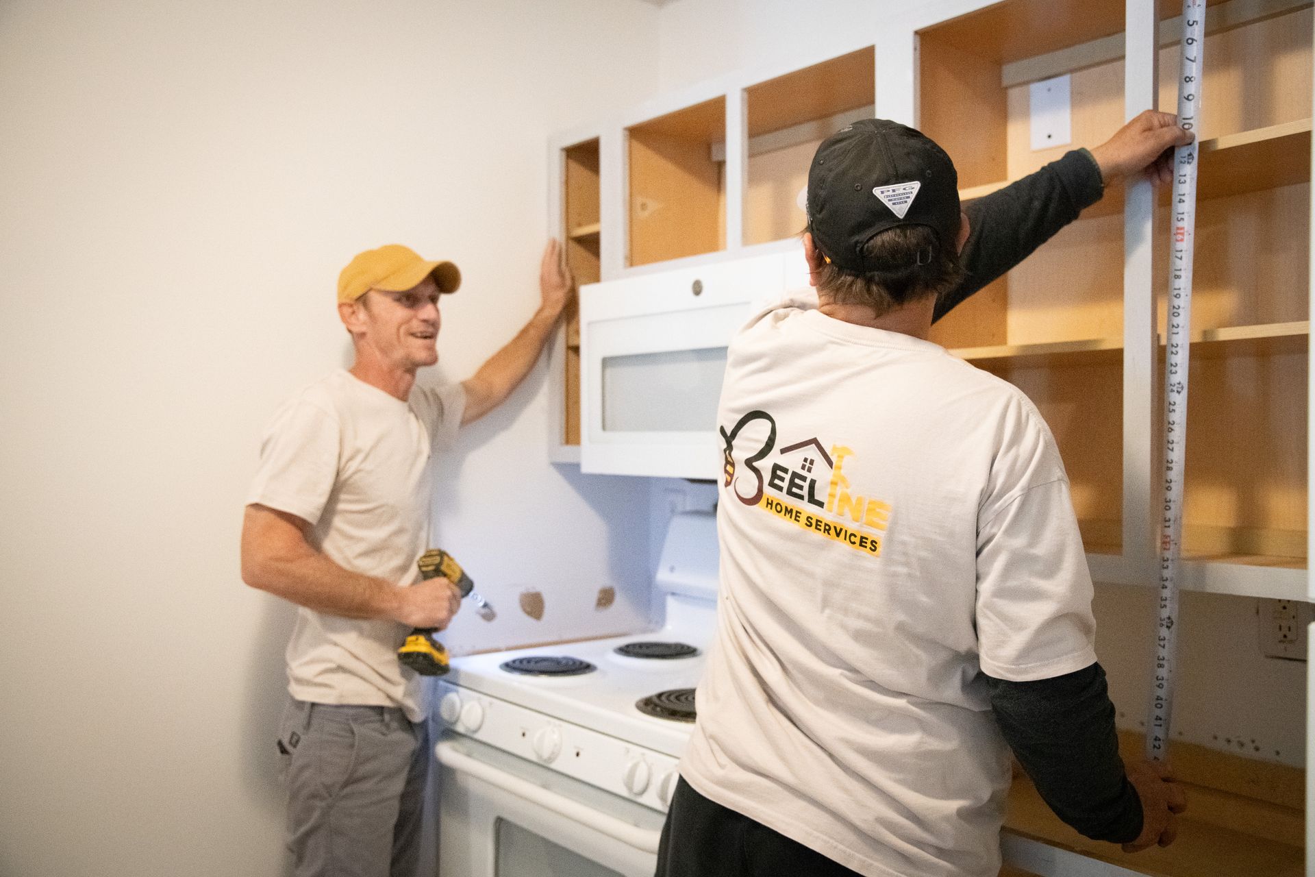 Two people installing kitchen cabinets over a stove. One holds a drill, the other measures with a ruler.