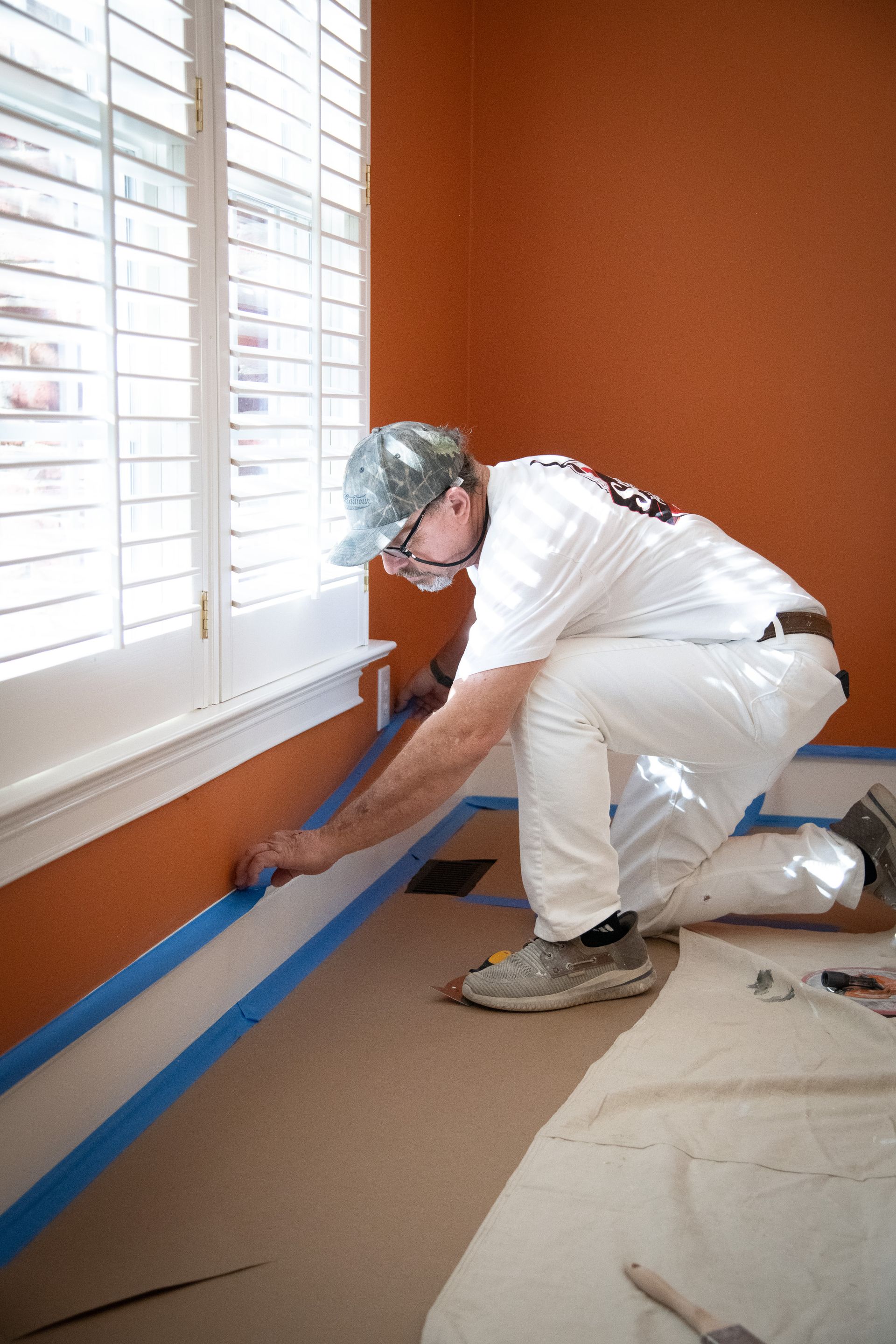 A person in white coveralls taping a baseboard for painting in an orange room.