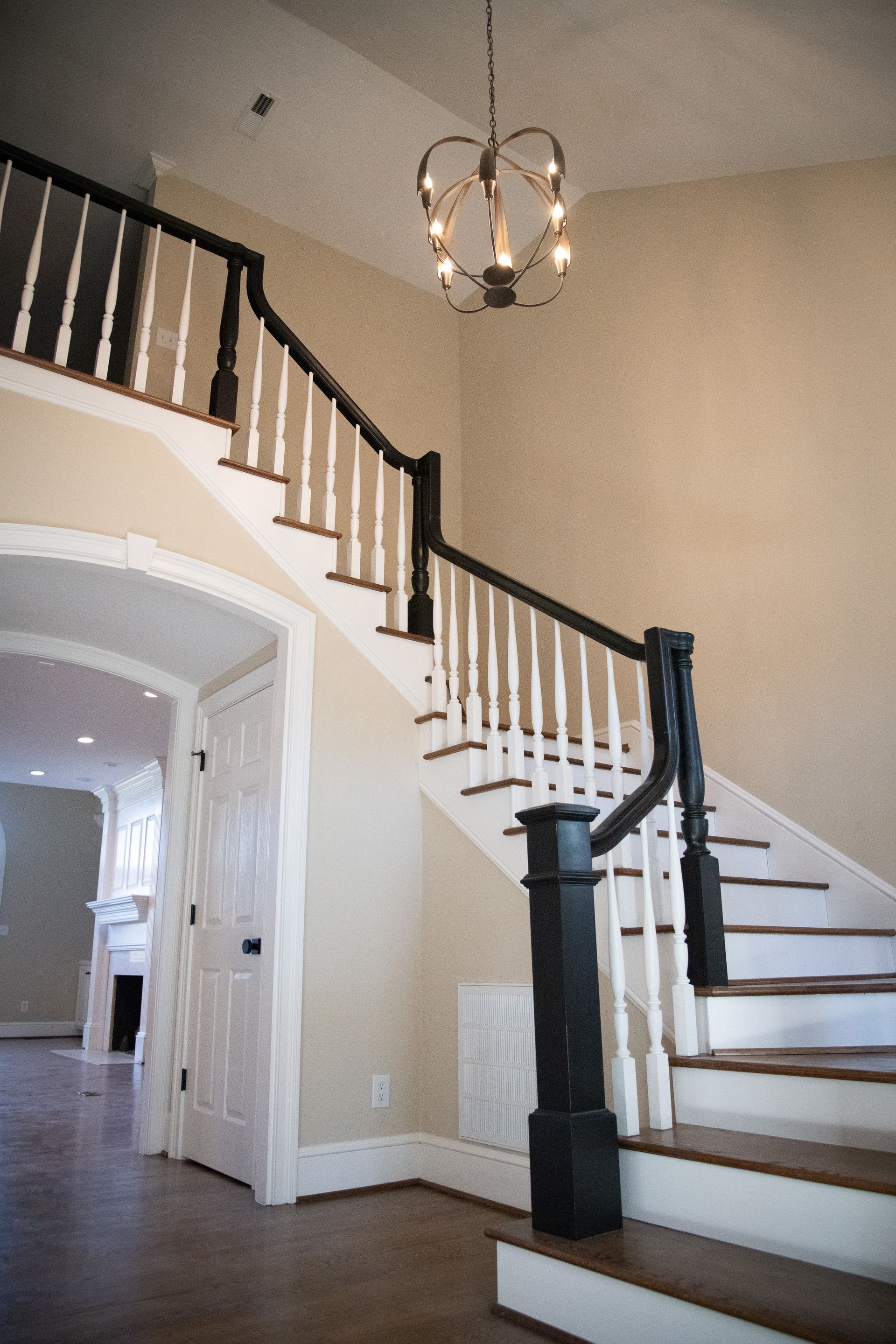 Staircase with black railing and white balusters, leading up to a chandelier in a neutral-toned foyer.