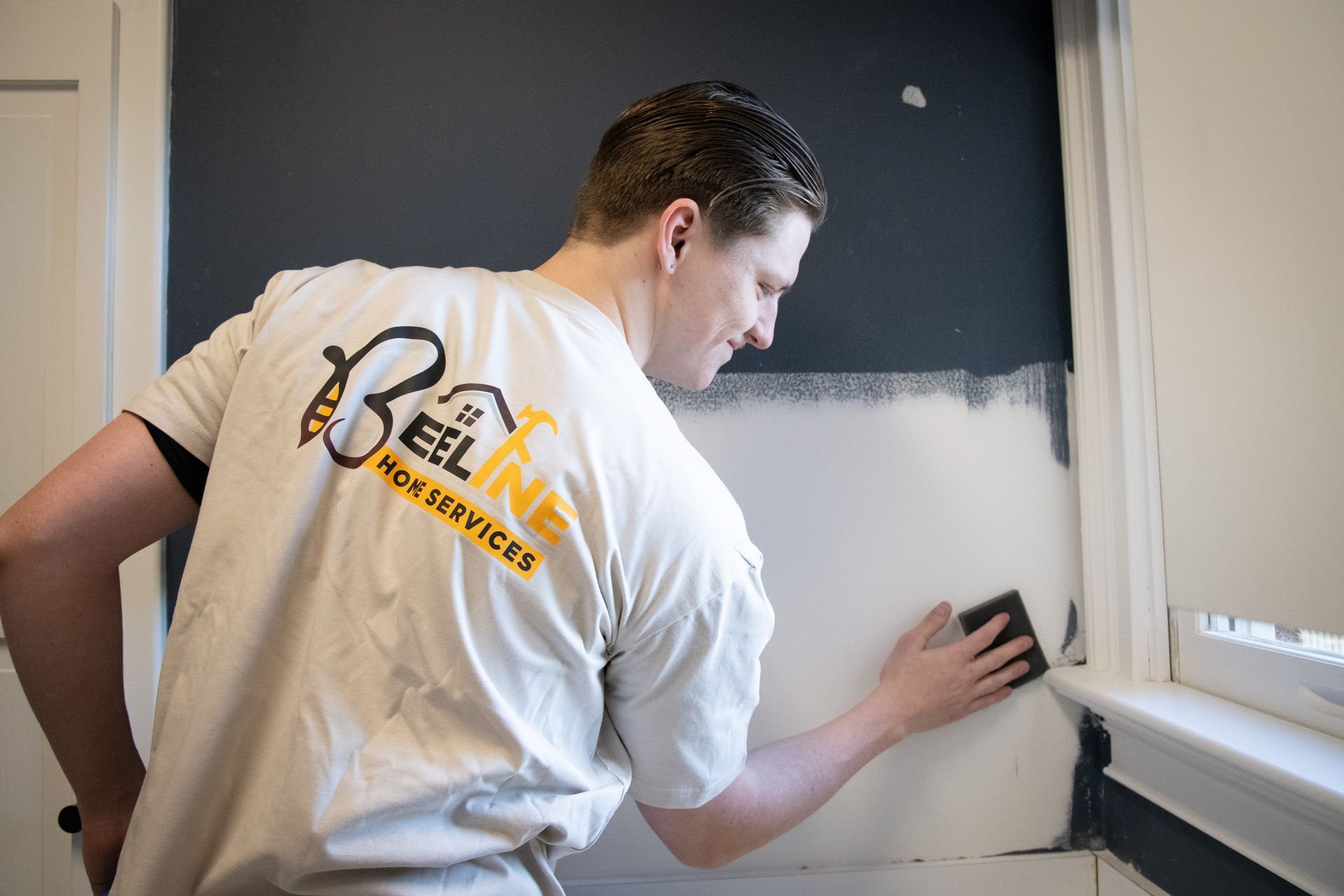 Person sanding a wall partially painted white, wearing a shirt with a company logo.
