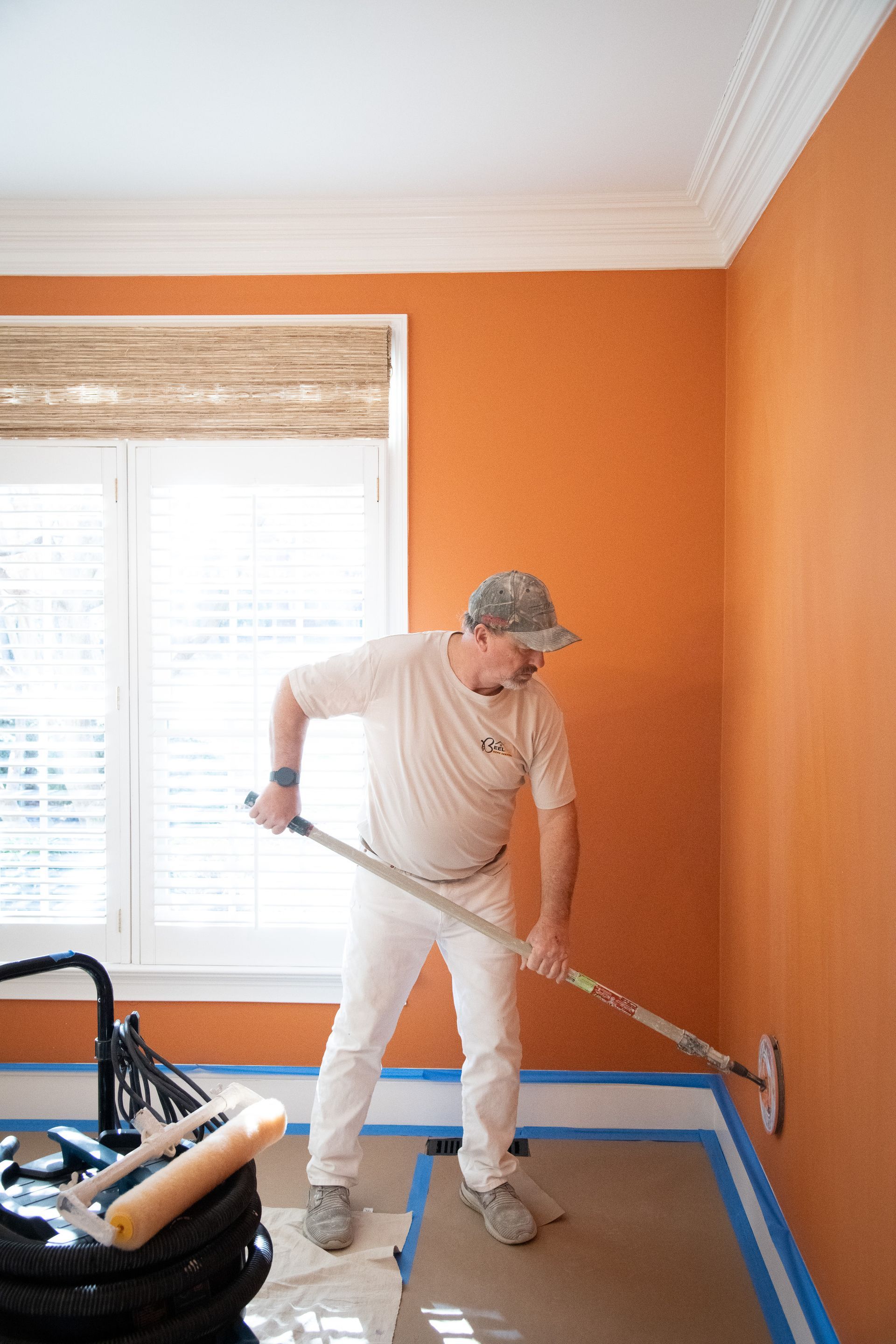 Person painting a wall orange indoors with a roller on a long handle, next to a window.