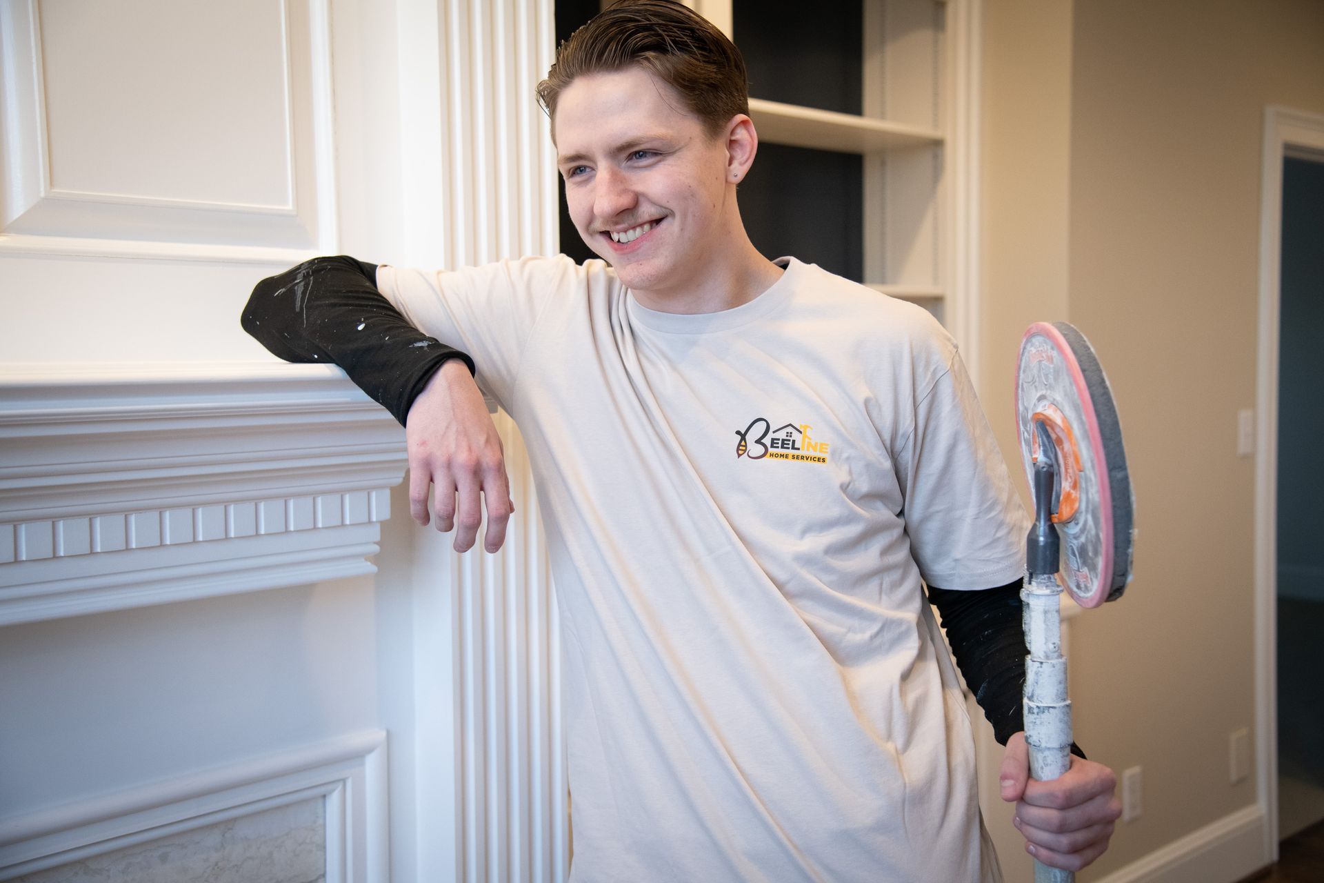 Man leans on mantel holding cleaning pad, smiling. Beige walls and cabinetry in background.