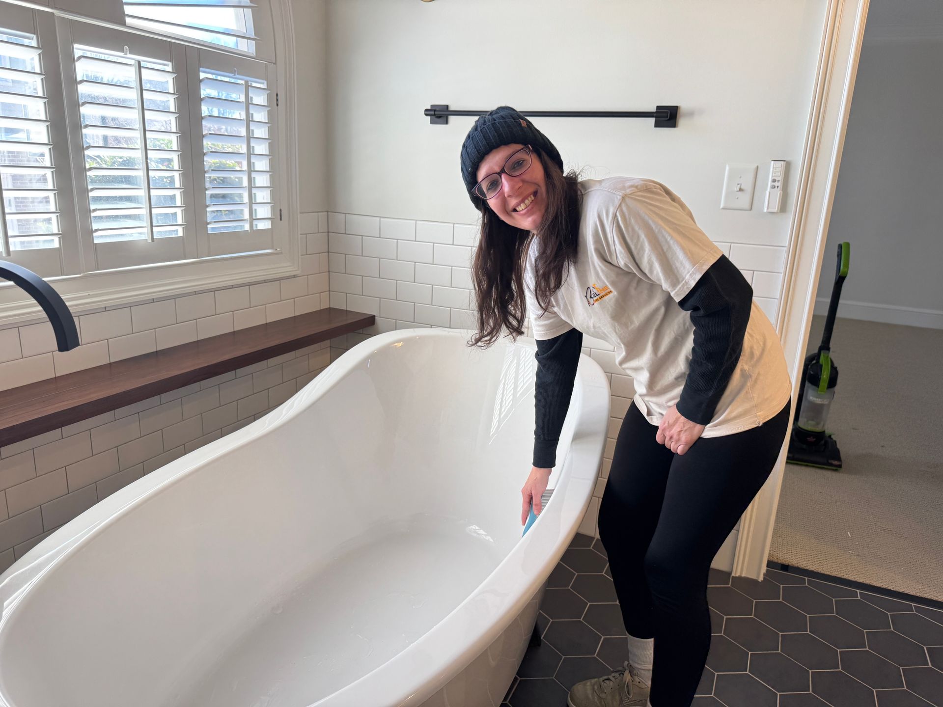 Woman in bathroom, cleaning white bathtub, smiling. Black beanie, black leggings, cream shirt.