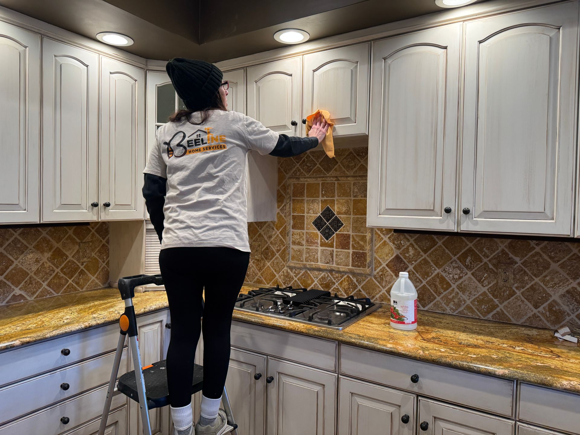 Person cleaning kitchen cabinets while standing on a small ladder.