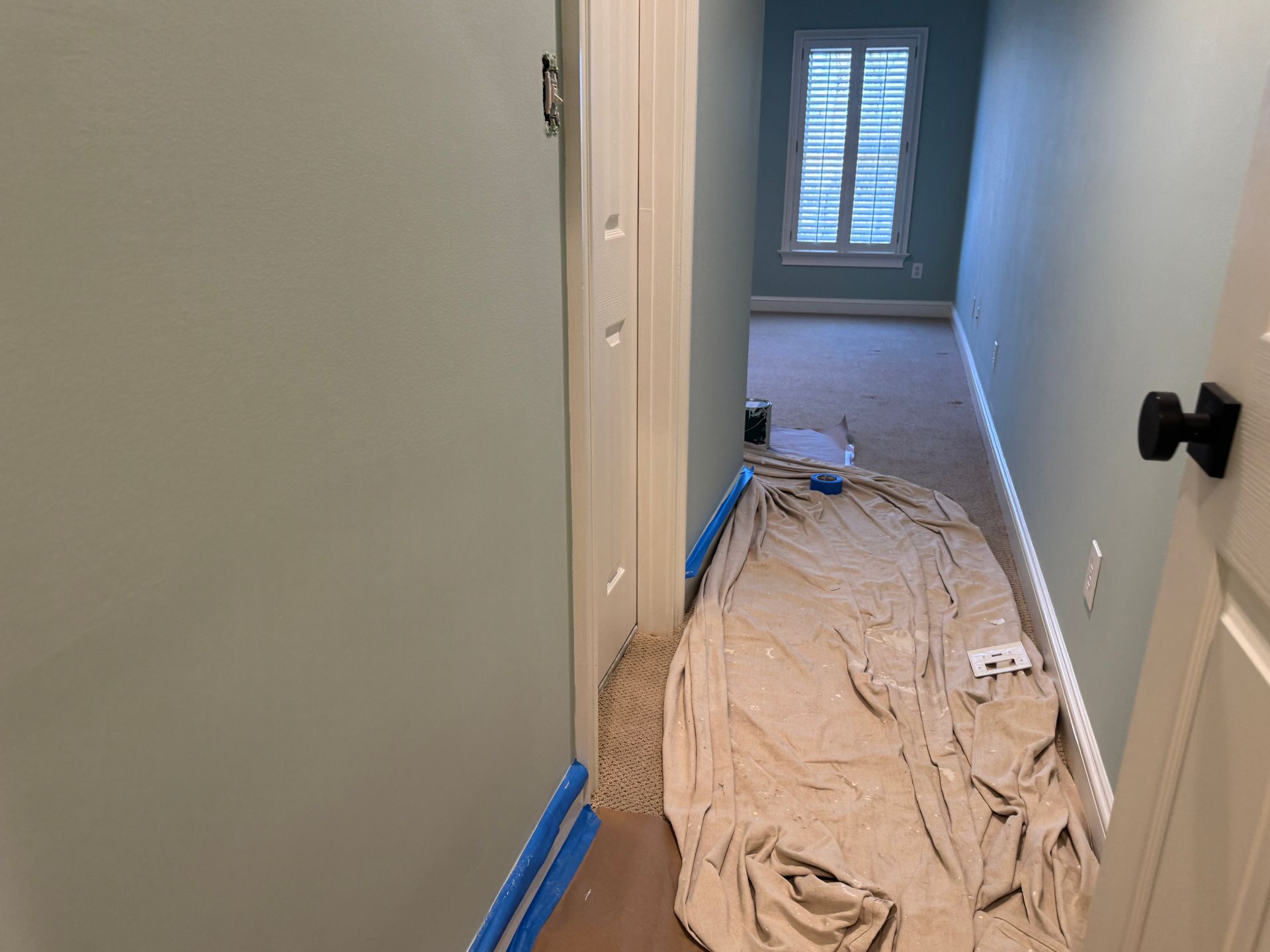 Hallway with blue-green walls leading to a room with a window; drop cloth on the floor.