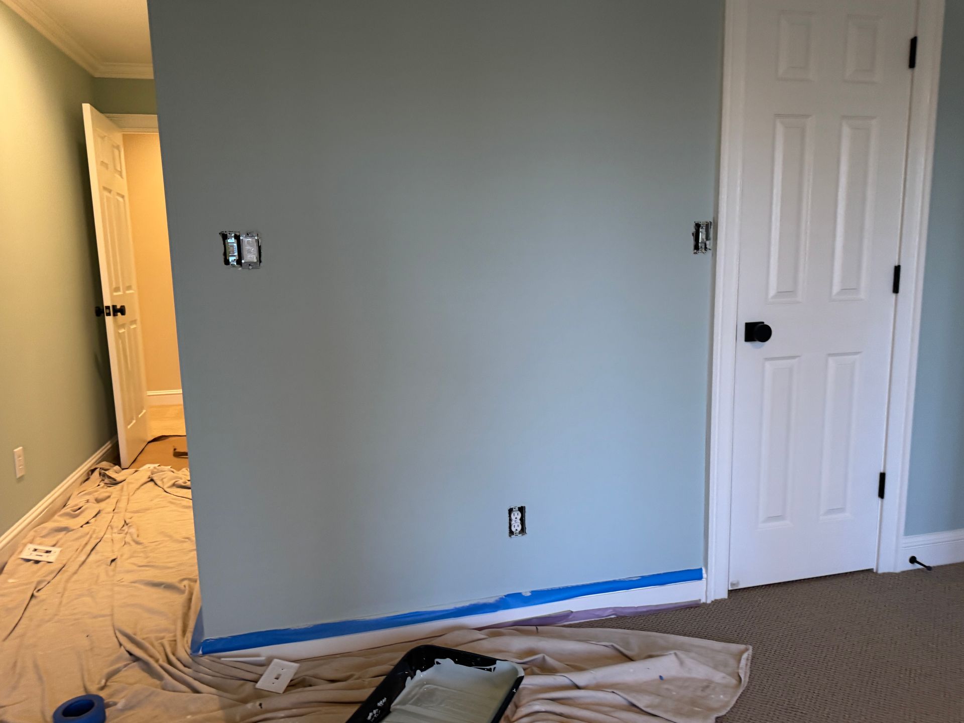 Room with light blue walls, white door, and blue painter's tape; paint supplies on a drop cloth.