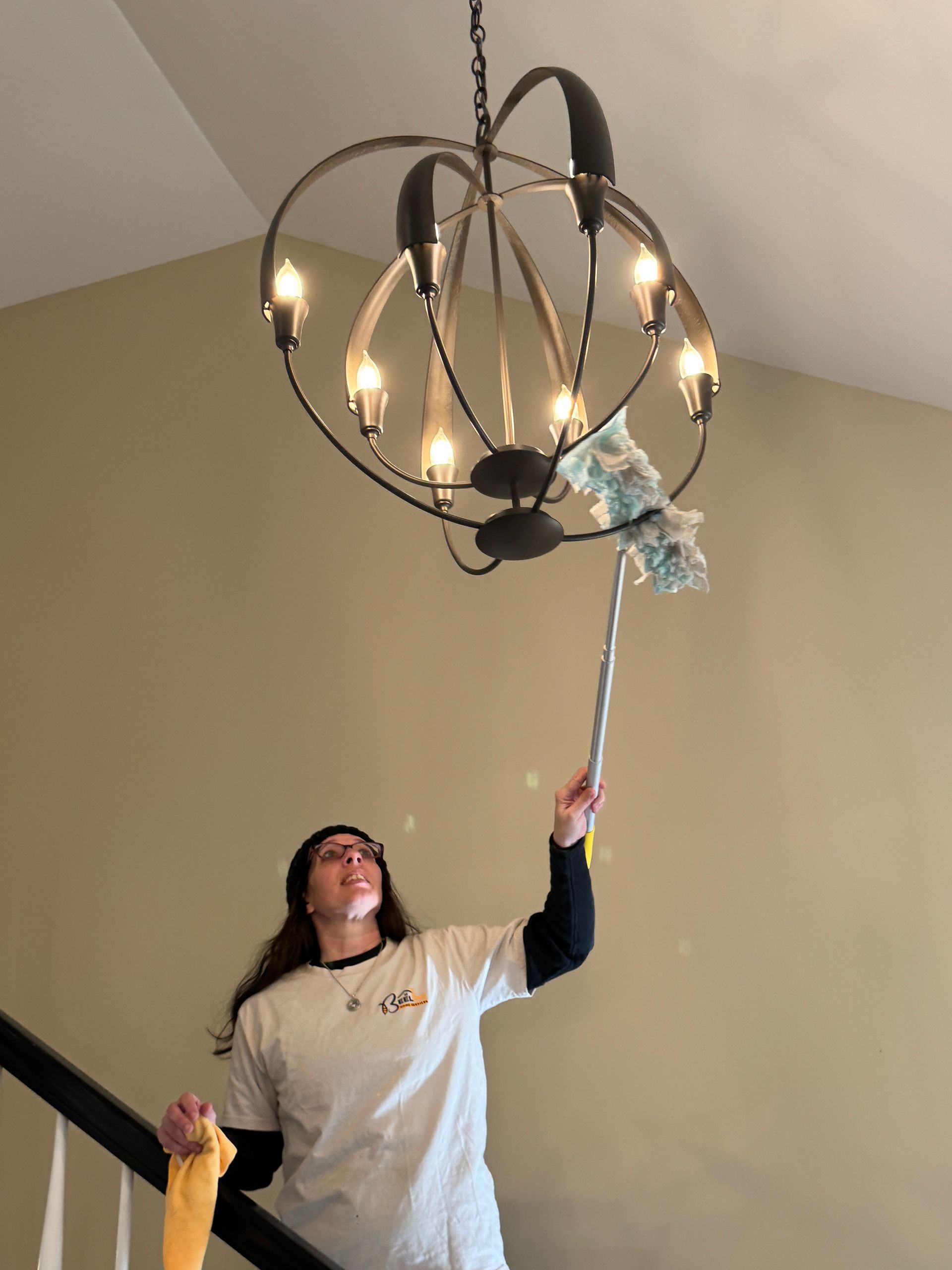 Person dusting a chandelier in a stairwell. They hold a duster in one hand, a cloth in the other.
