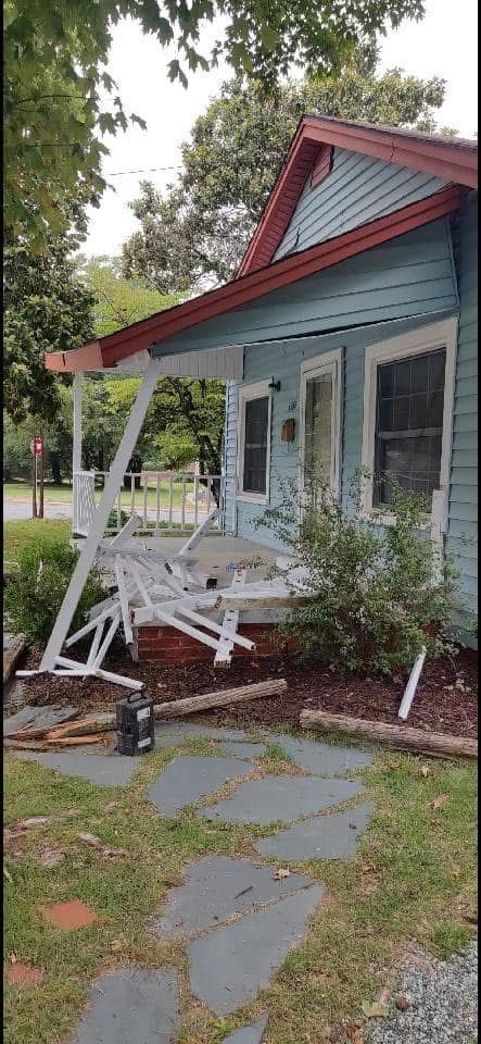 A blue house with a collapsed porch swing sits among trees.