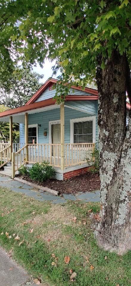 Light blue cottage with porch, under a leafy tree, in front of a green lawn.