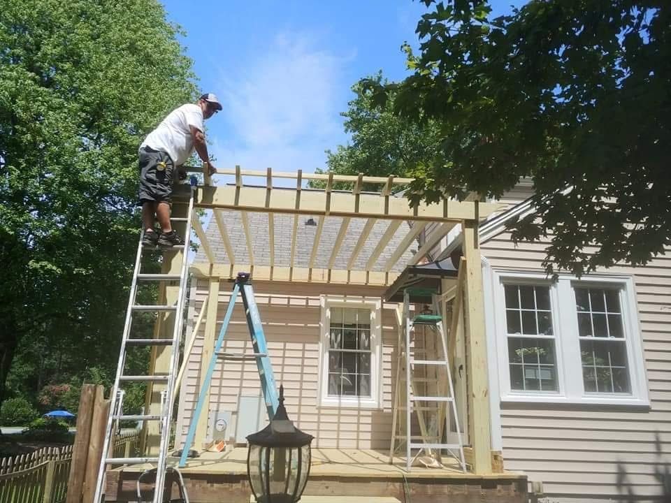 Person on ladder constructing wooden pergola over a porch. Sunny outdoor setting with a house.