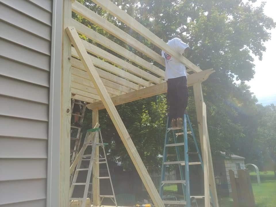 Person on ladder building a wooden pergola attached to a house; sunny outdoor setting.