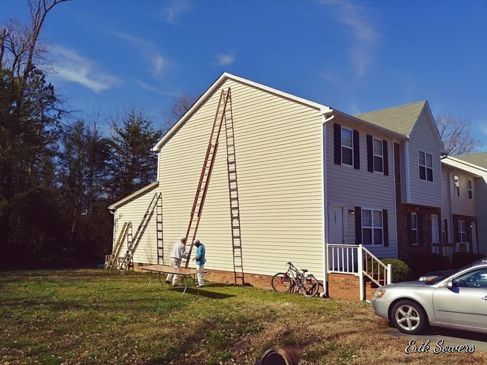 Workers painting a two-story building's siding, using tall ladders. A car and bicycles sit nearby, sunny day.