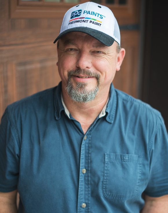 Man in a blue shirt and baseball cap smiles outdoors.  Door in background.