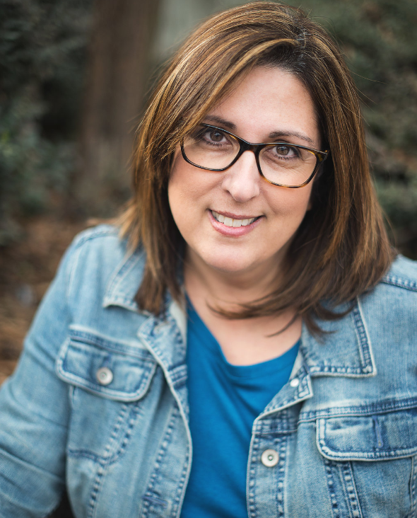 Woman with glasses smiles outdoors, wearing a denim jacket and blue shirt.