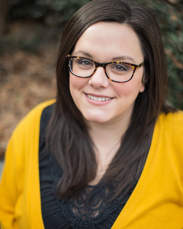 Woman with glasses, smiling, wearing a yellow sweater and black shirt, outdoors with trees in the background.