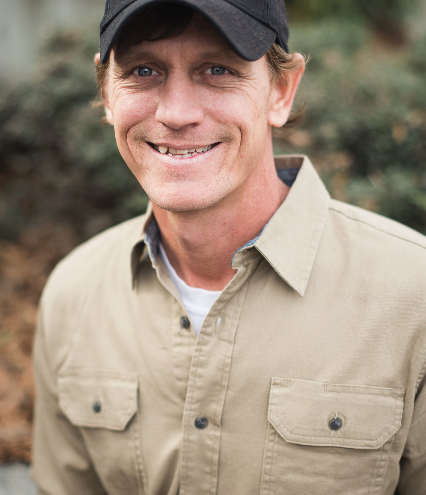 Man wearing a black cap and khaki shirt smiles outdoors with blurred greenery in the background.