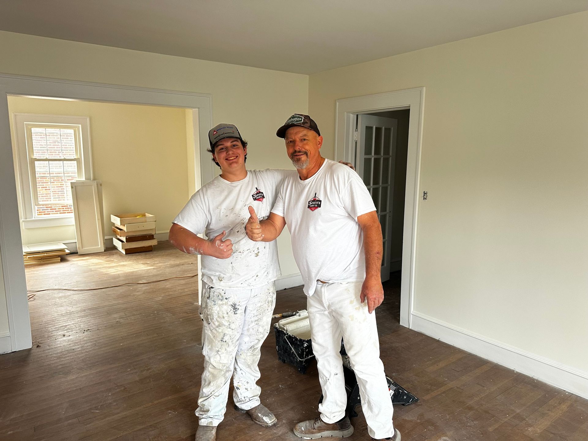 Two people in paint-splattered white clothes inside a room with freshly painted light yellow walls. One gives a thumbs up.