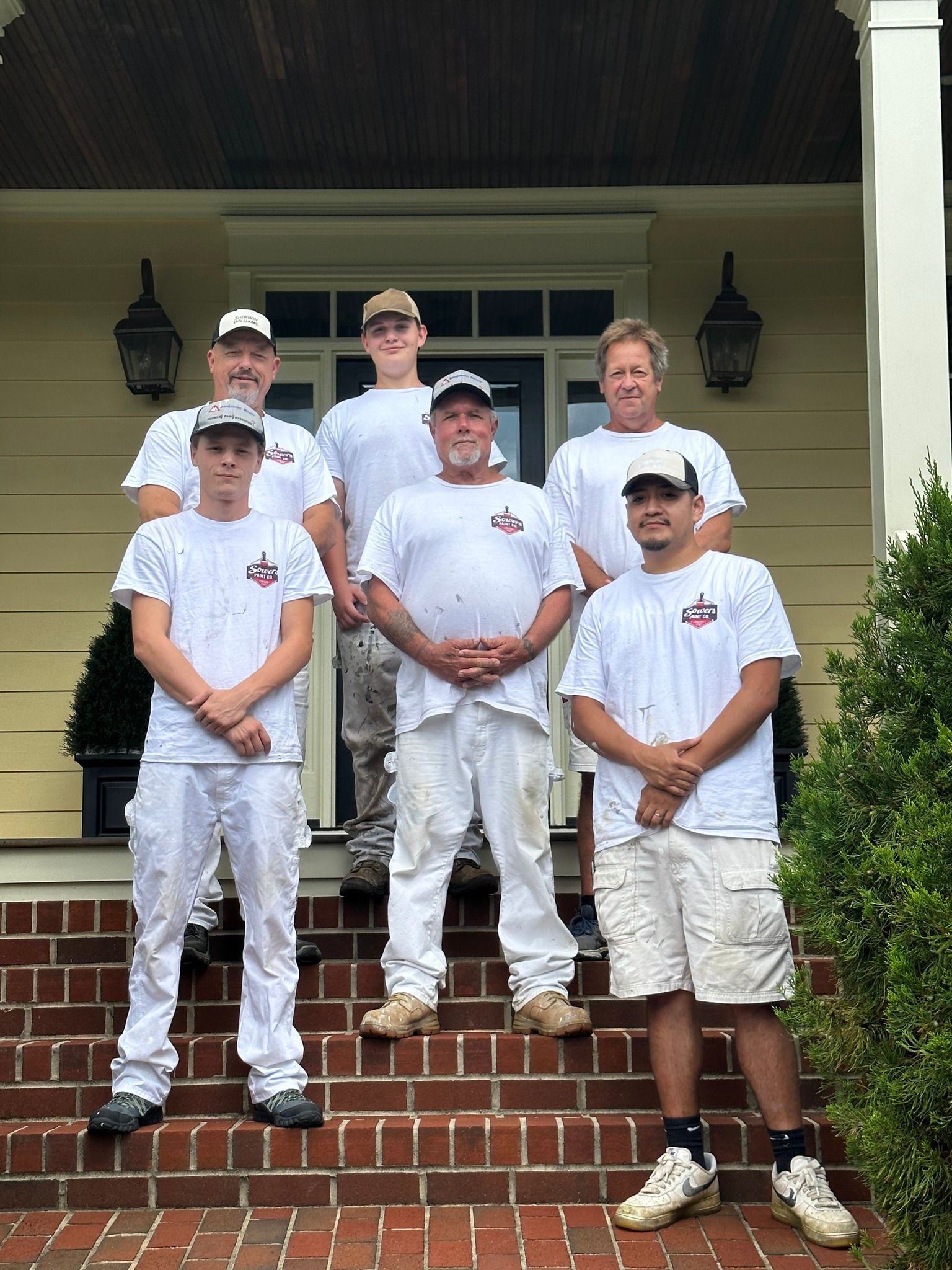 Group of painters posing on brick steps in front of a house. All wear white shirts and pants, some with paint stains.