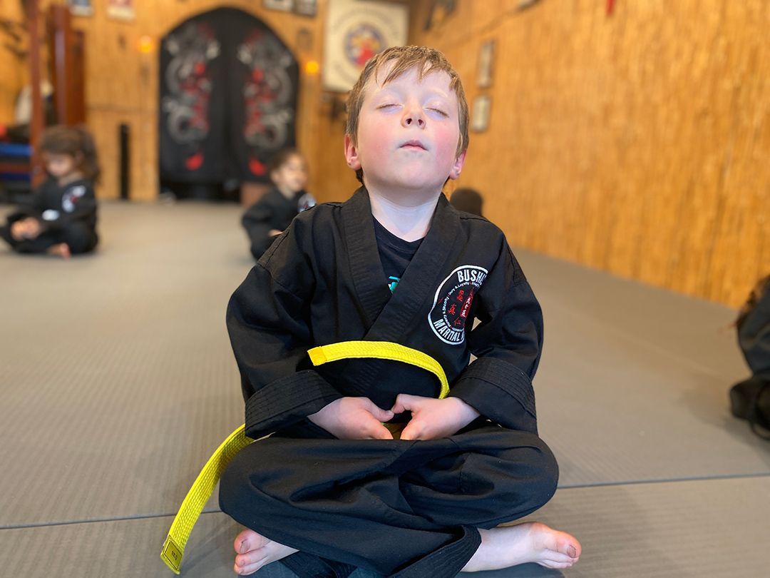 A young boy in a black karate uniform is sitting in a lotus position with his eyes closed.
