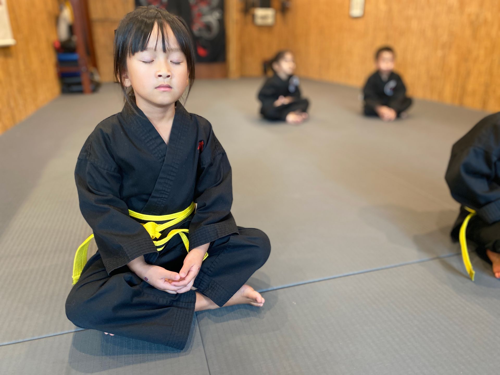 A young girl in a black karate uniform is sitting in a lotus position on a mat.