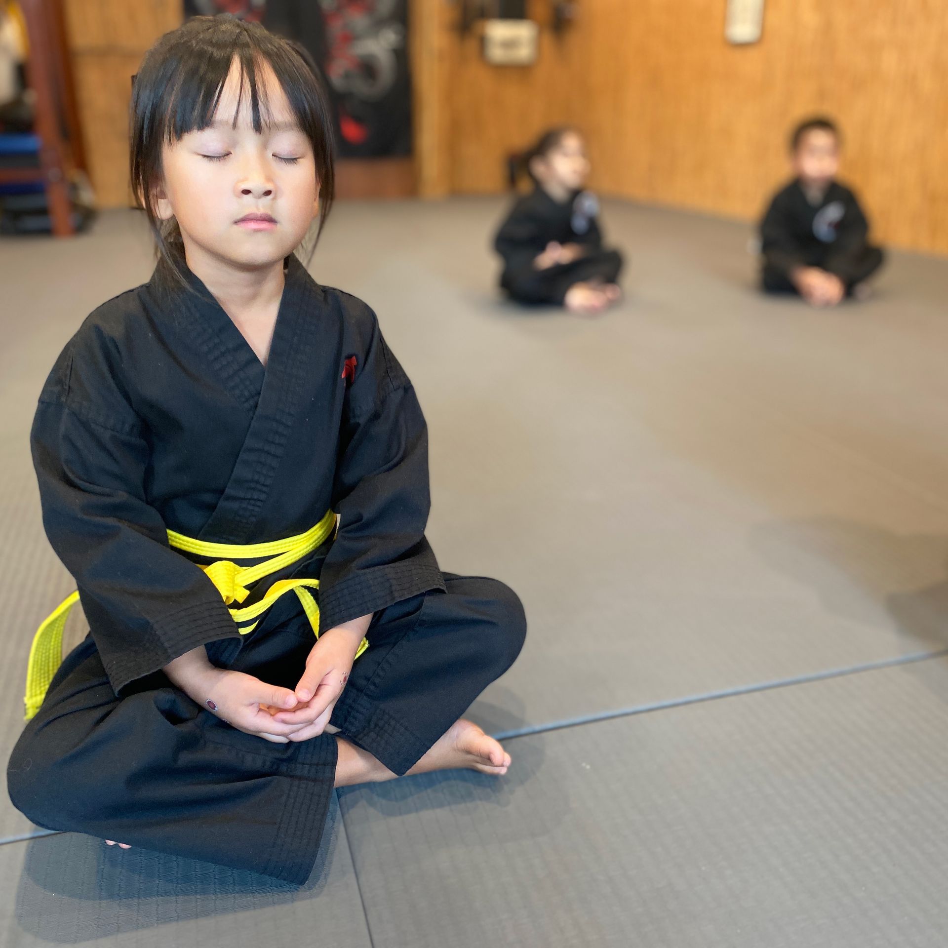 A little girl in a black karate uniform sits on the floor with her eyes closed