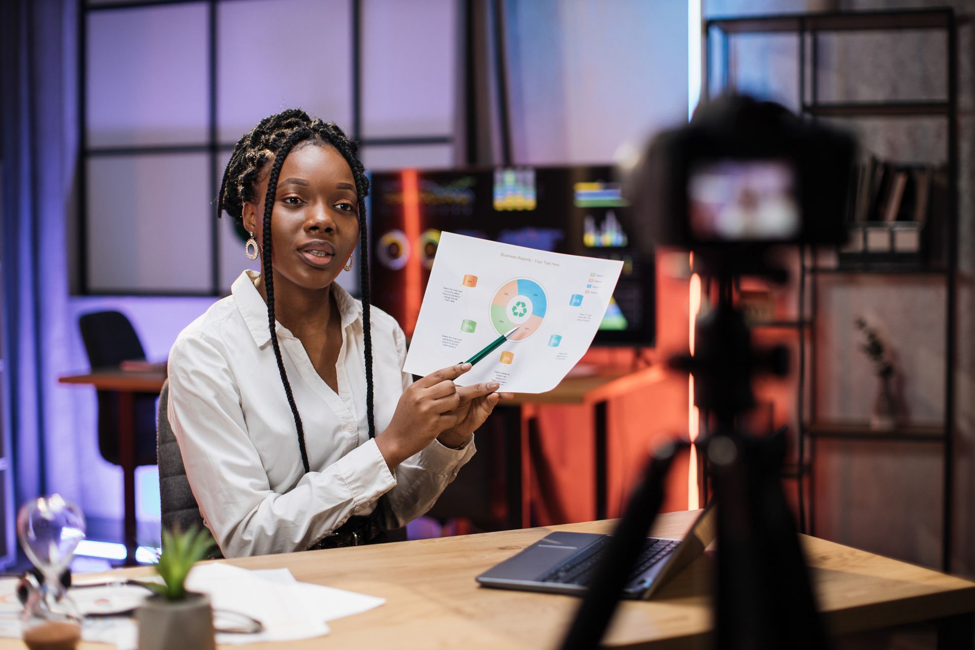 A woman is sitting at a desk holding a piece of paper in front of a camera.