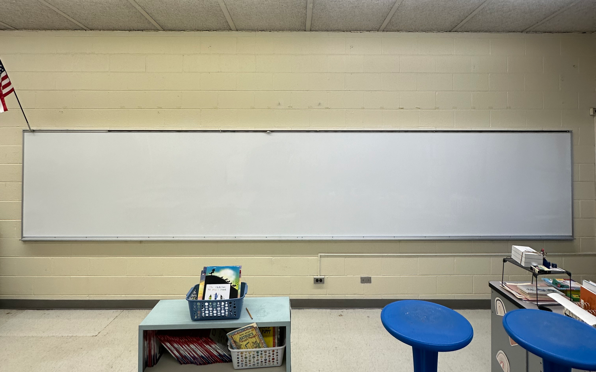 Classroom whiteboard on beige wall with blue stools and a small supply cart below