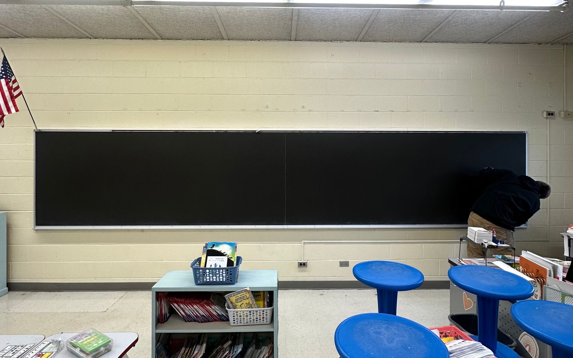 Empty classroom with a long black chalkboard, blue stools, and a bookshelf beneath it.