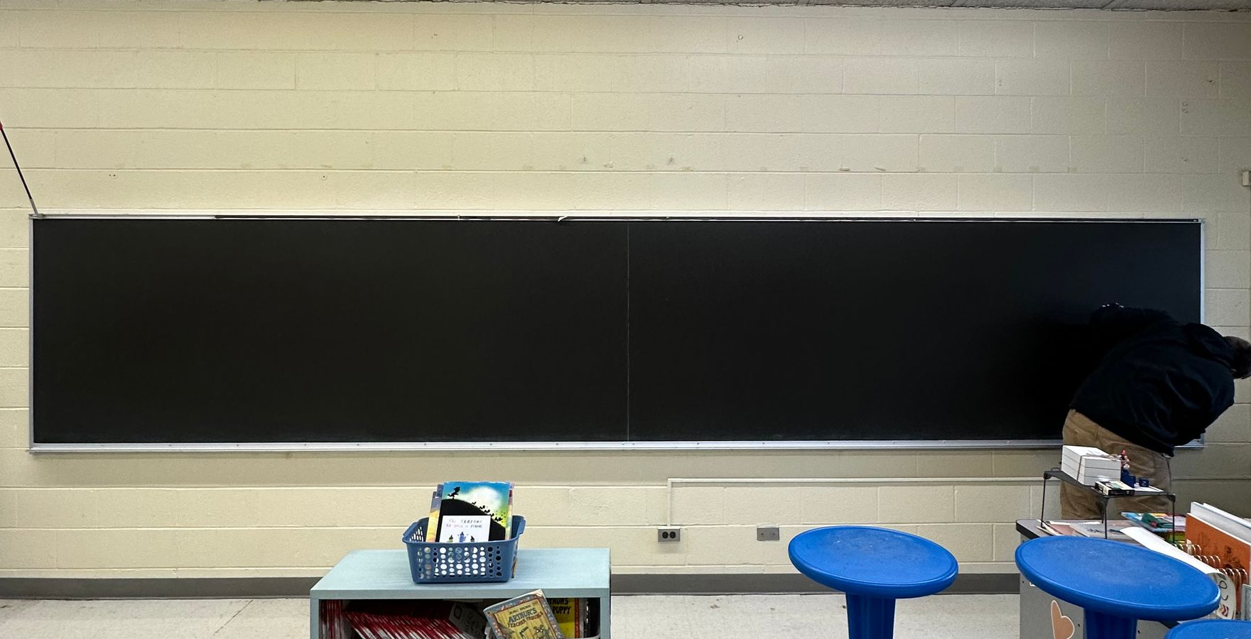 Classroom with a long blackboard, small table, and blue stools under a beige wall.