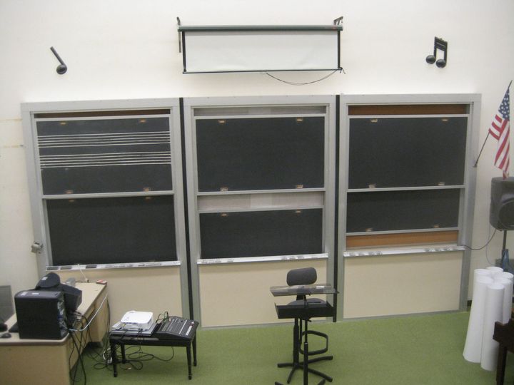 Empty classroom with three windows, music notes on the wall, and a music stand in front of green carpet.